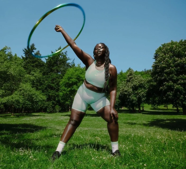 Athletic woman outdoors in workout gear, hula hooping in park on sunny day. Trees and blue sky in background.