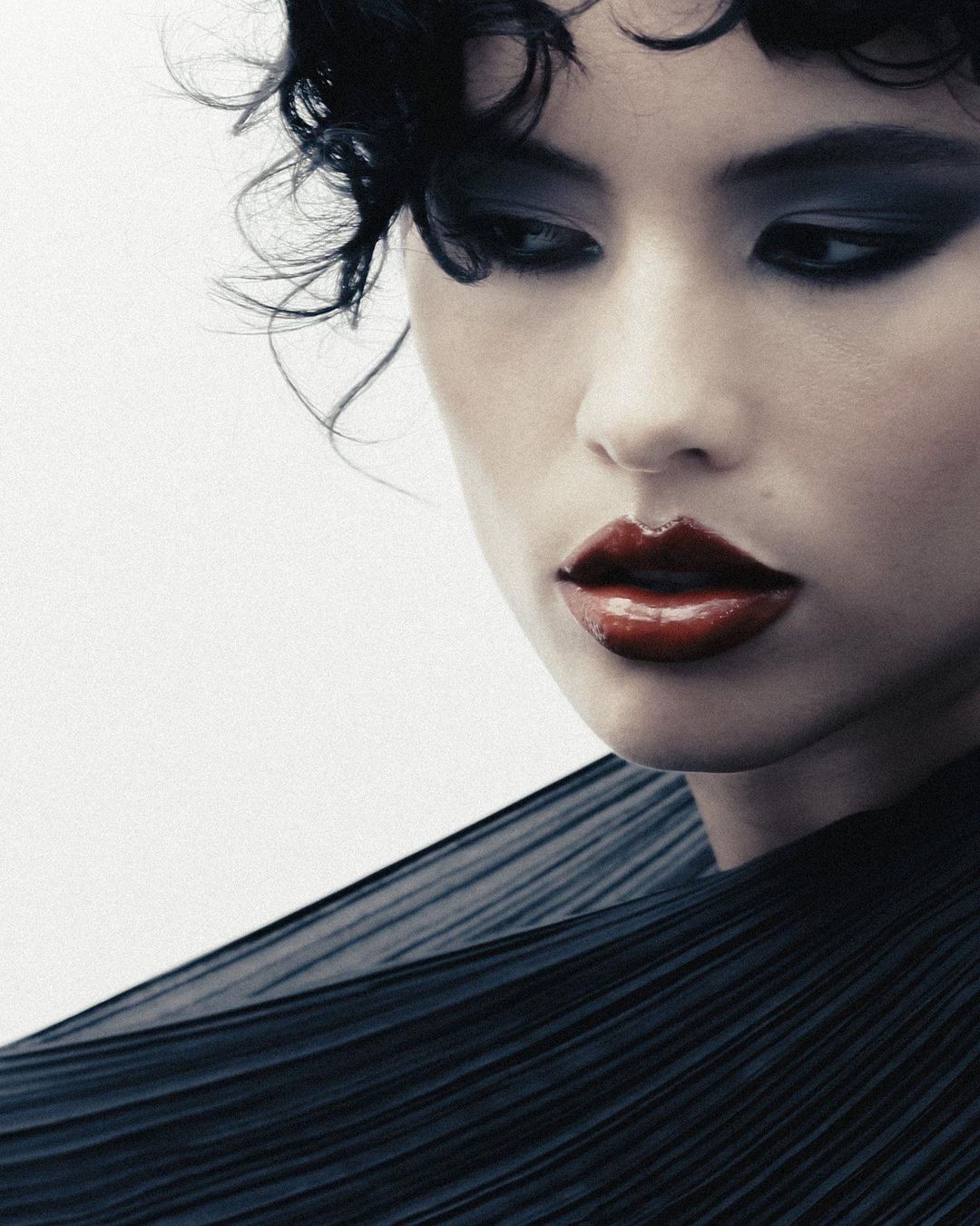 Close-up portrait of a woman with dark curly hair, smoky eye makeup, and glossy red lips, wearing a dark pleated top against a white background.
