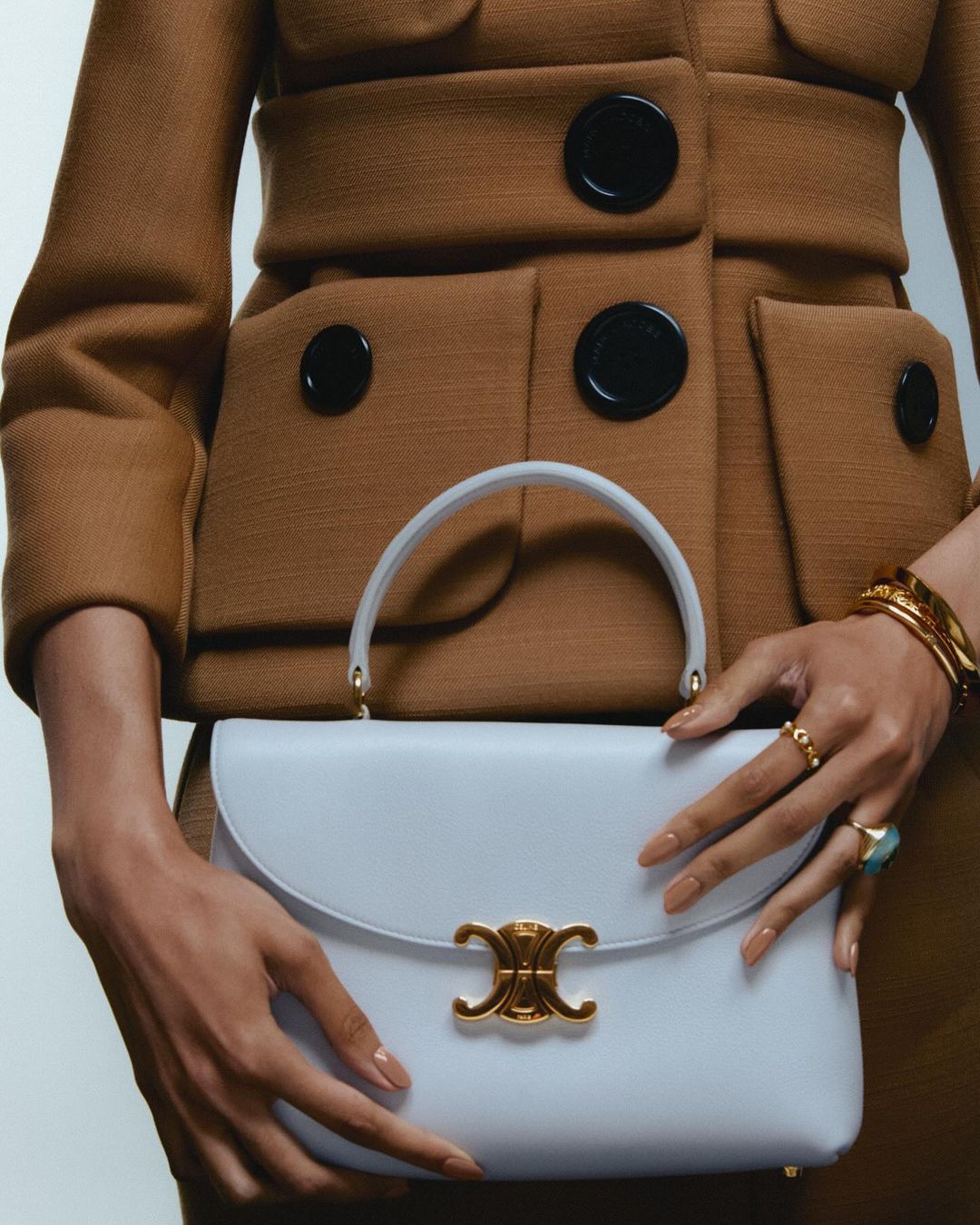 Woman in brown jacket holds a blue leather handbag with gold clasp. Close-up shot features gold bracelets and rings, studio lighting.