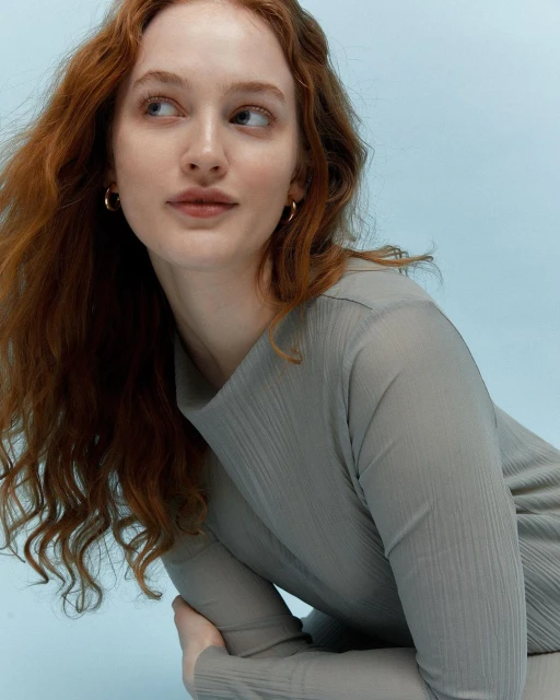 Close-up portrait of a woman with long red hair and a neutral expression against a light blue background, wearing a gray textured top.