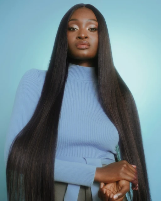 A confident dark-skinned woman poses in a light blue turtleneck and long sleek hair against a gradient blue backdrop in a studio shot.