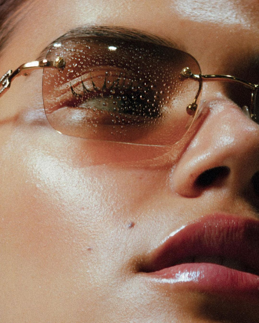 Close-up portrait of a person wearing tinted rimless sunglasses covered in water droplets with glossy lips