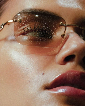 Close-up portrait of a person wearing tinted rimless sunglasses covered in water droplets with glossy lips