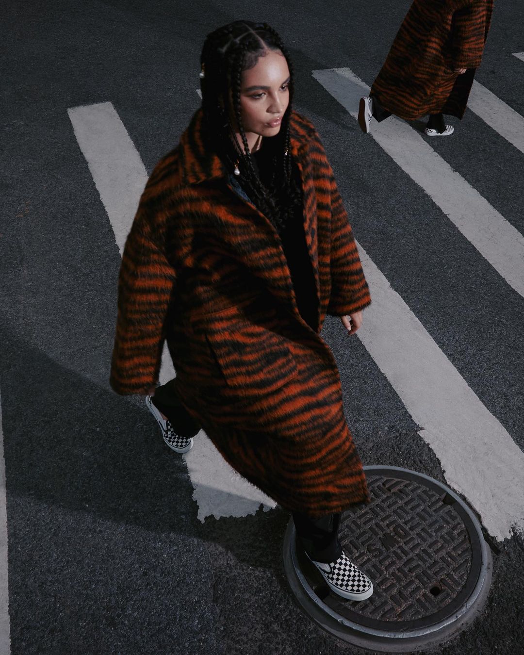 Woman in tiger-striped coat walks on crosswalk in urban setting. Vans shoes, overhead shot, dark mood.