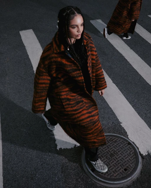 Woman in tiger-striped coat walks on crosswalk in urban setting. Vans shoes, overhead shot, dark mood.
