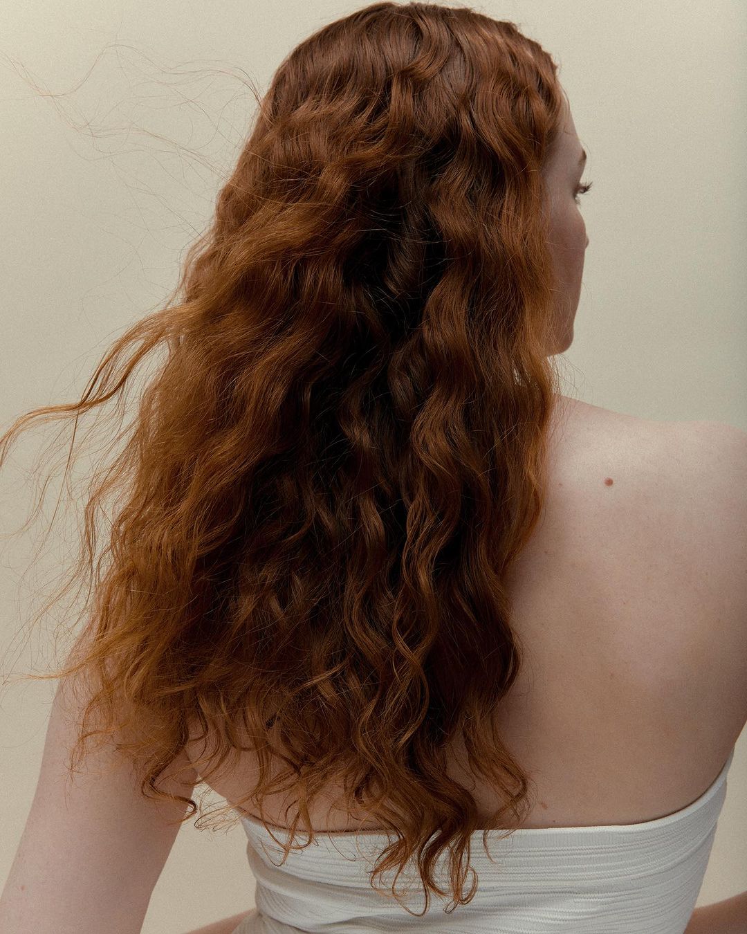 Rear view of a woman with long wavy red hair wearing a white strapless top against a seamless cream background.