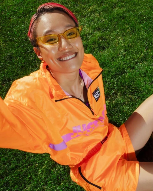 Smiling person in an orange Nike windbreaker and shorts sitting on grass, wearing yellow glasses, looking at the camera outdoors.