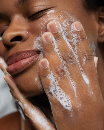 Close-up of a woman with dark skin washing her face with a foamy cleanser, eyes closed, showing beauty and skincare routine.