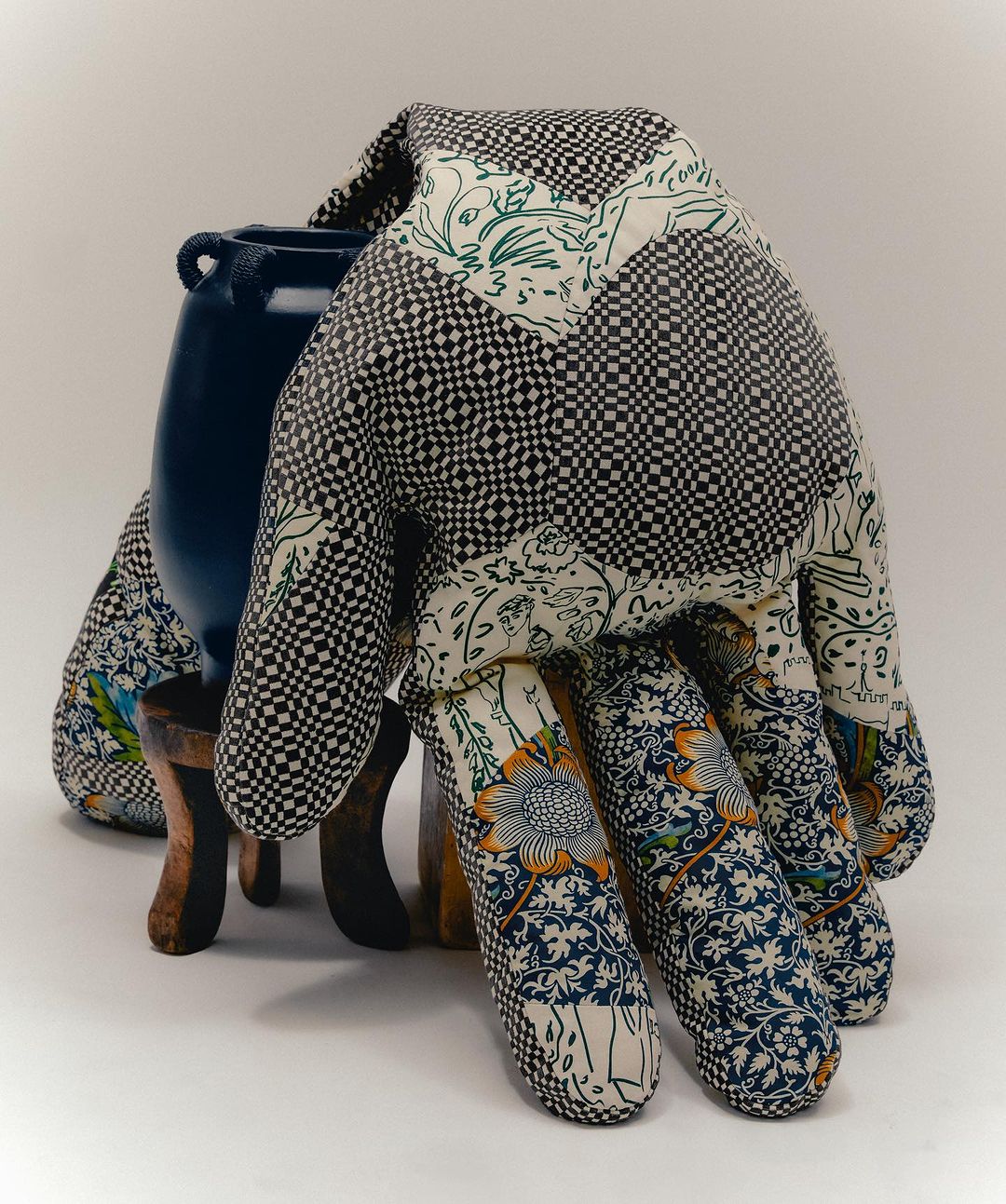 Still life of a blue ceramic vase atop a brown wooden stool, draped with a patterned, quilted textile in a studio setting.