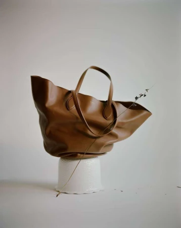 Brown leather tote bag resting on a white textured stand against a plain neutral background with a delicate twig.