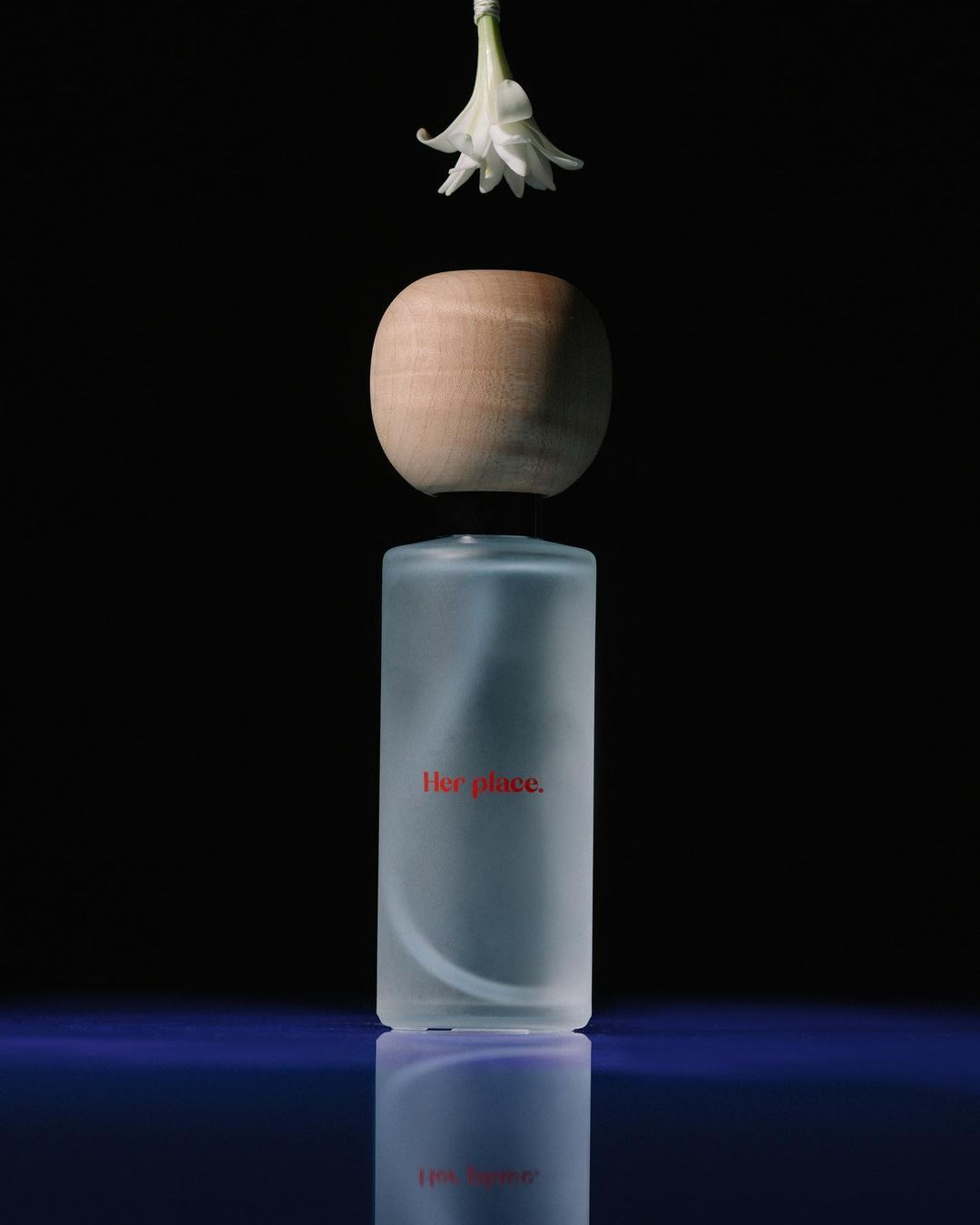 A minimalist still life featuring a cylindrical frosted glass bottle, a wooden spherical cap, and a white flower against a black background.