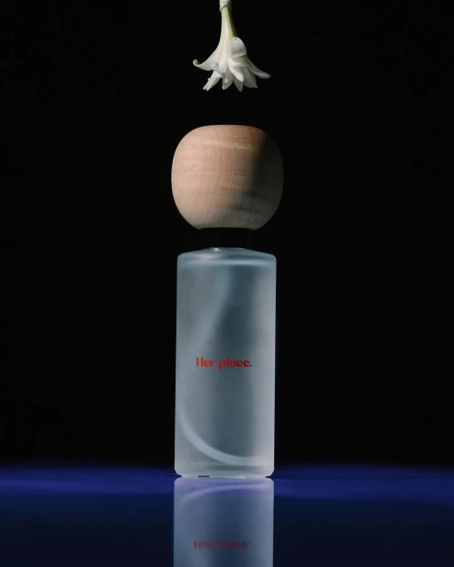 A minimalist still life featuring a cylindrical frosted glass bottle, a wooden spherical cap, and a white flower against a black background.