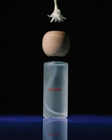 A minimalist still life featuring a cylindrical frosted glass bottle, a wooden spherical cap, and a white flower against a black background.