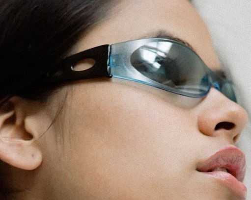 Close-up of a woman's face wearing reflective blue-framed futuristic sunglasses in a side-profile studio shot.
