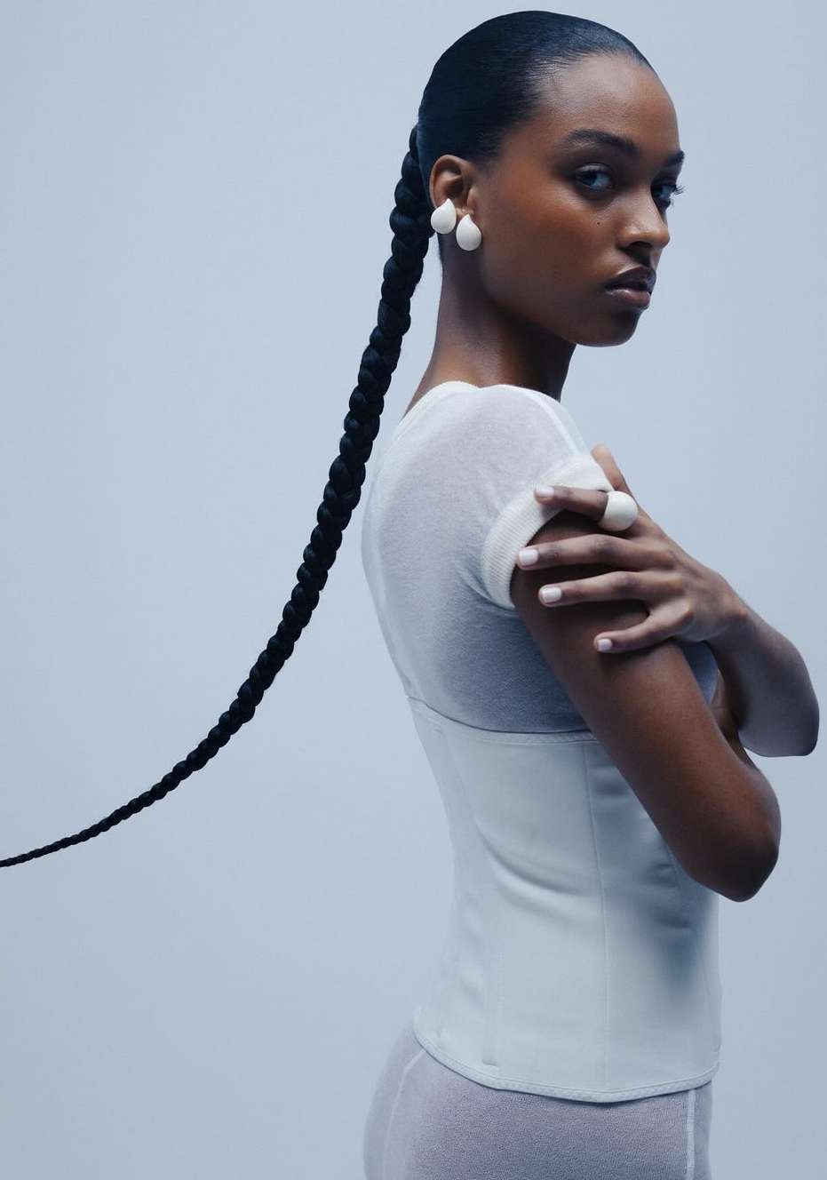 Portrait of a Black woman with long braided hair, wearing white earrings, a white top, and a ring, against a light blue background.