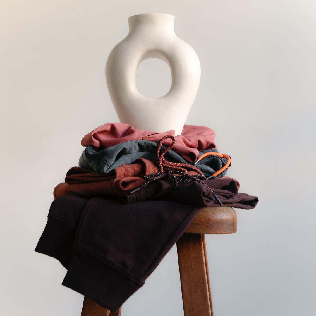 Still life of a stack of apparel in mauve, brown, grey, and black on a wooden stool, topped with a cream-colored modern vase.