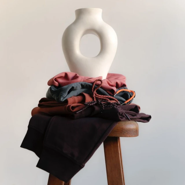Still life of a stack of apparel in mauve, brown, grey, and black on a wooden stool, topped with a cream-colored modern vase.