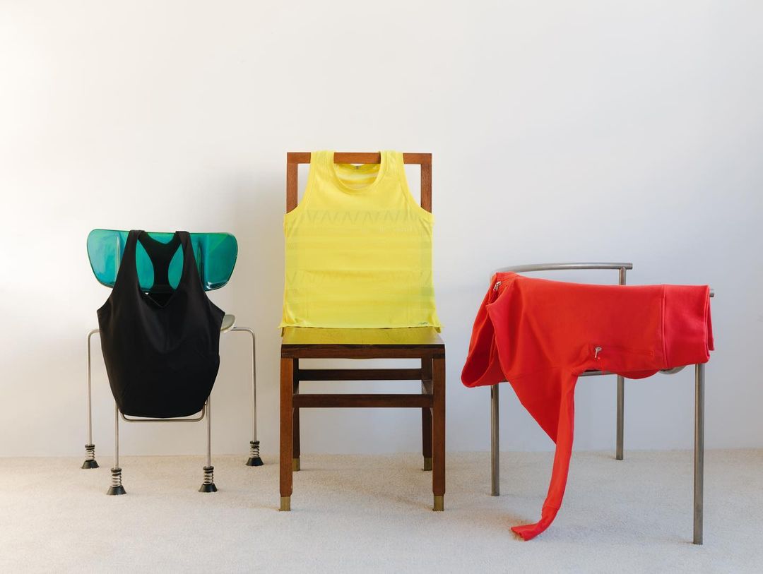 Three chairs each draped with clothing: a black tank top, yellow tank top, and a red zip-up hoodie; minimalist studio shot.