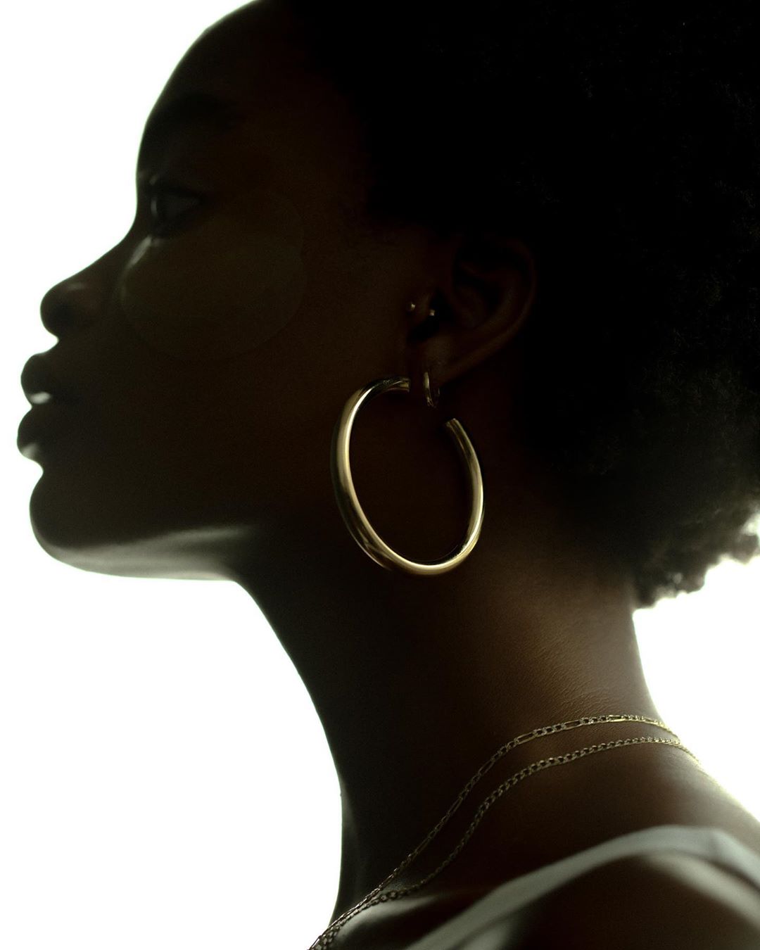 Close-up side profile of a dark-skinned woman with gold hoop earrings and necklace against a bright white background.