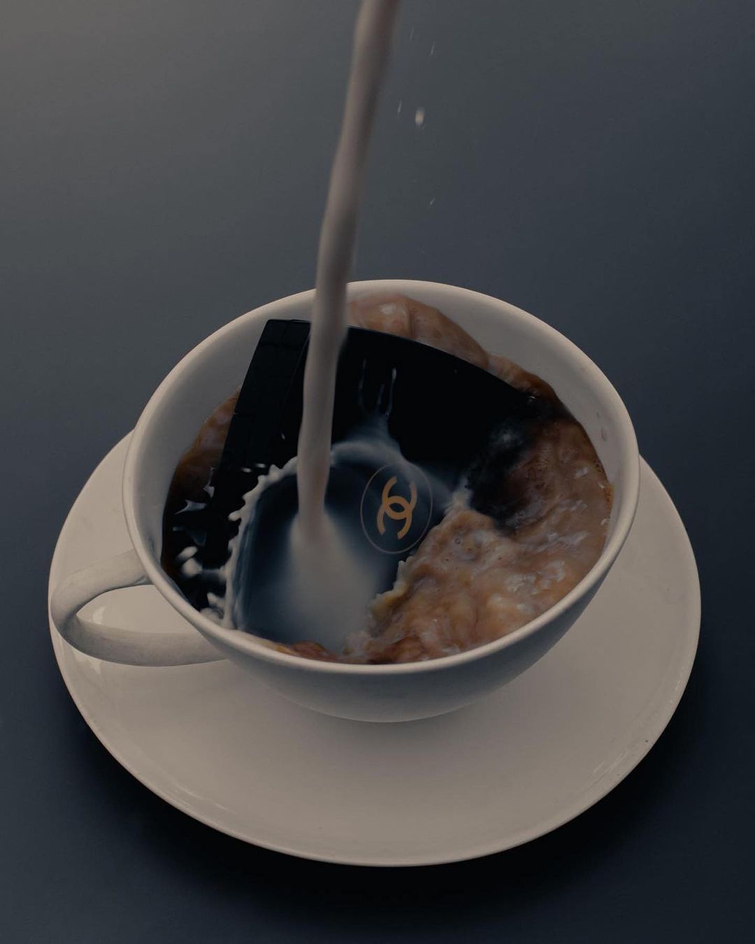 Milk being poured into a white ceramic teacup filled with cereal; a Chanel logo is visible beneath the stream of liquid.