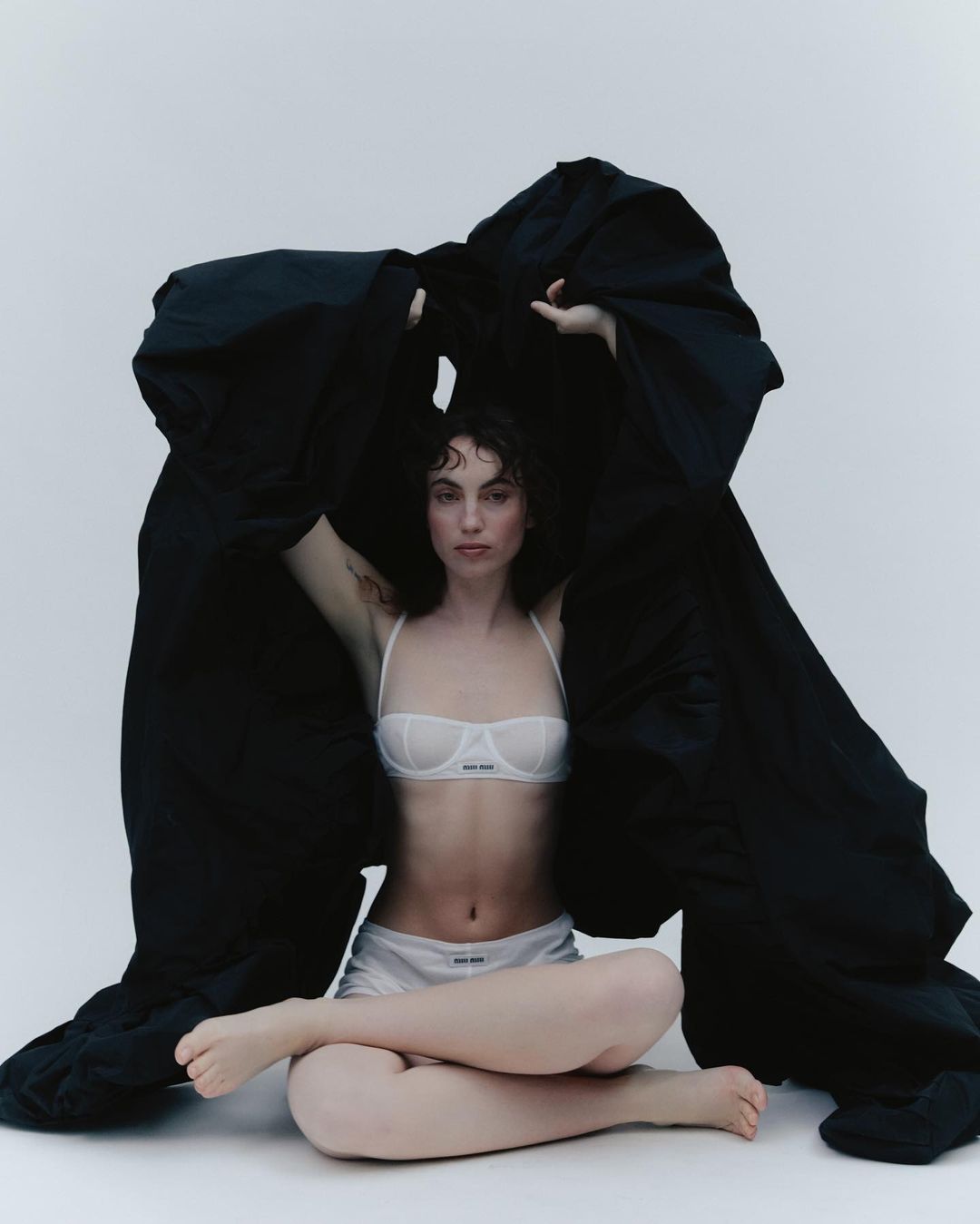 A young woman in lingerie sits cross-legged beneath a draped black cloth in a fashion pose against a plain white backdrop.