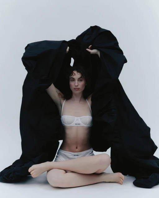 A young woman in lingerie sits cross-legged beneath a draped black cloth in a fashion pose against a plain white backdrop.