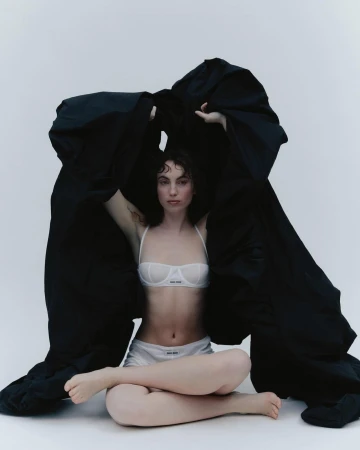 A young woman in lingerie sits cross-legged beneath a draped black cloth in a fashion pose against a plain white backdrop.