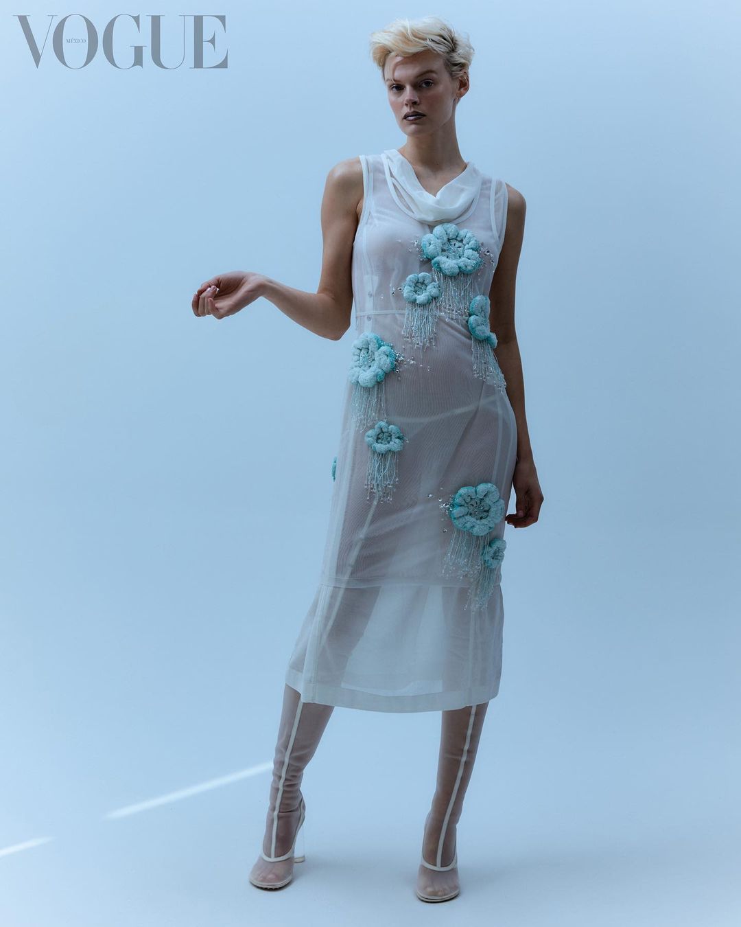 A slender model poses in a sheer dress with pale blue floral appliqués in a studio shot for Vogue, wearing sheer beige heeled booties.
