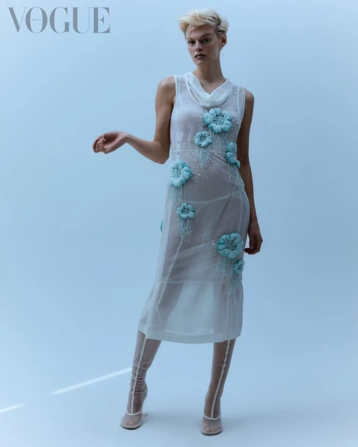 A slender model poses in a sheer dress with pale blue floral appliqués in a studio shot for Vogue, wearing sheer beige heeled booties.