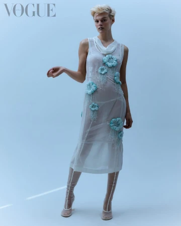 A slender model poses in a sheer dress with pale blue floral appliqués in a studio shot for Vogue, wearing sheer beige heeled booties.