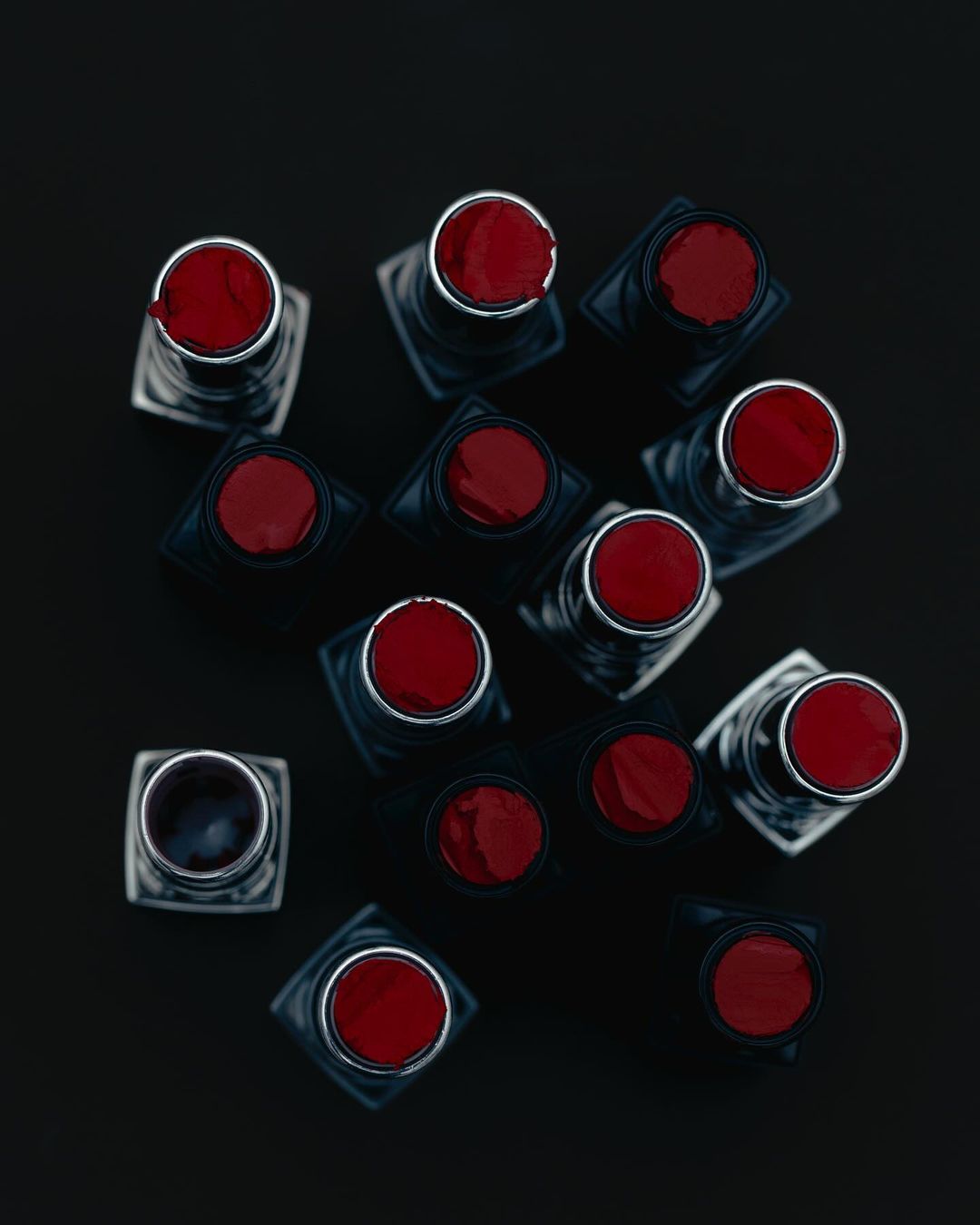 Overhead shot shows a collection of red lipsticks with black bases, arranged on a stark black background, creating a bold, modern beauty image.