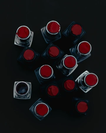 Overhead shot shows a collection of red lipsticks with black bases, arranged on a stark black background, creating a bold, modern beauty image.