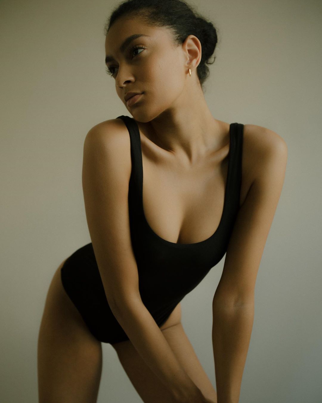 Portrait of a young, dark-haired woman in a black bodysuit looking off camera in a studio setting with a neutral background and soft lighting.