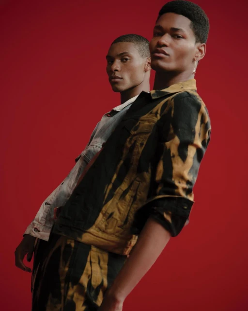Two adult men pose in a fashion shot against a red backdrop, wearing jackets, facing the camera in a studio with soft, diffused lighting.