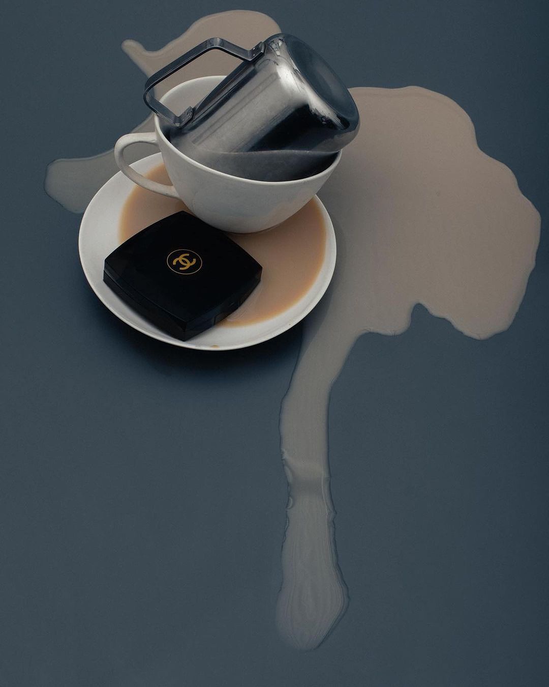 Overhead shot of a spilled coffee cup on a gray surface with a black compact makeup case nearby, creating a minimalist, still-life composition.