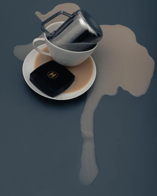 Overhead shot of a spilled coffee cup on a gray surface with a black compact makeup case nearby, creating a minimalist, still-life composition.