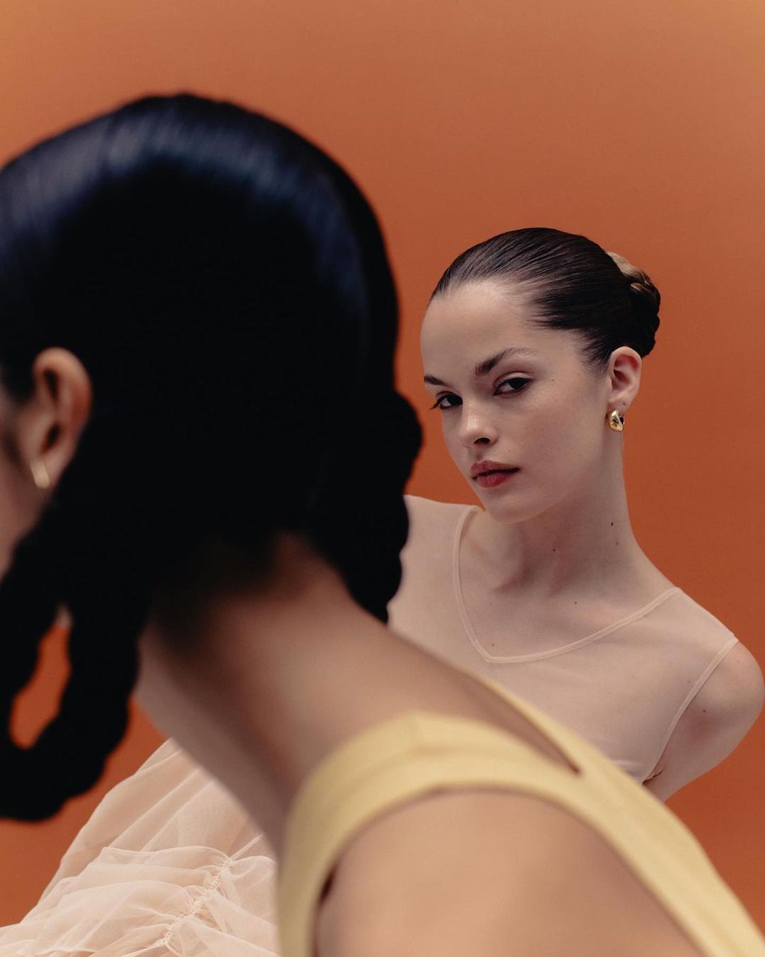 Two women pose against a bright orange background. One woman looks at the camera, wearing a sheer top. The other is facing away.