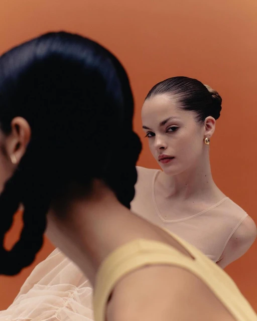 Two women pose against a bright orange background. One woman looks at the camera, wearing a sheer top. The other is facing away.