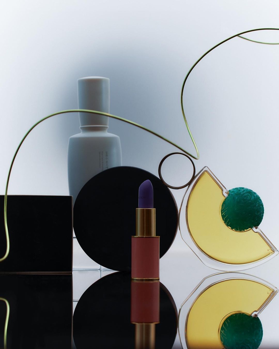 Still life of purple lipstick, white perfume bottle, and black and yellow geometric shapes against a gradient white background. 