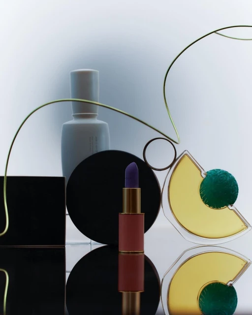 Still life of purple lipstick, white perfume bottle, and black and yellow geometric shapes against a gradient white background. 