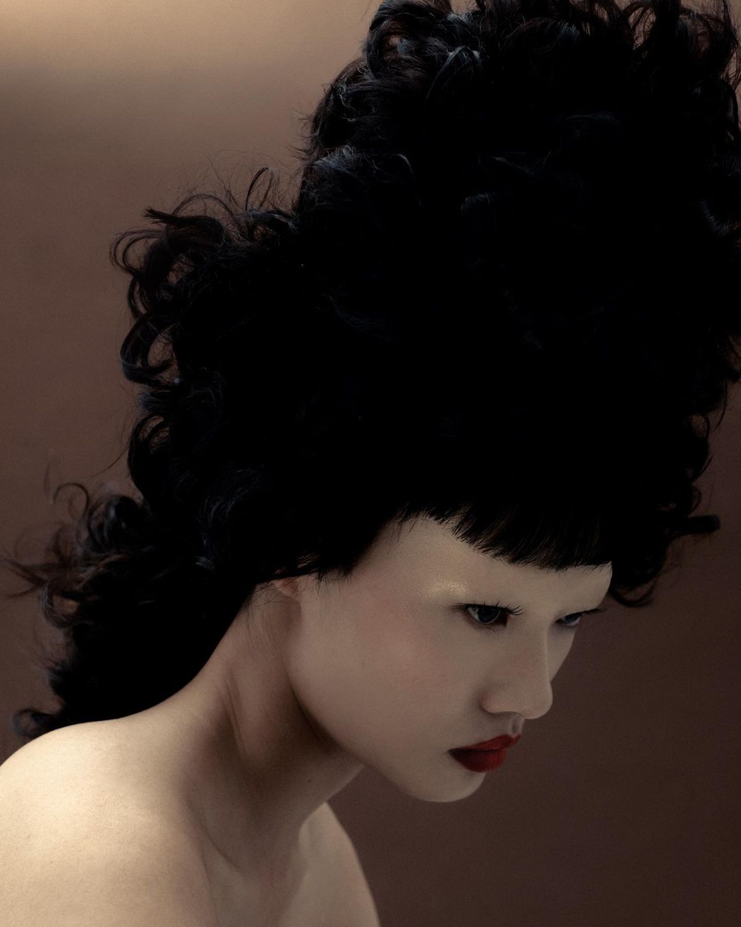 Studio shot of a fashionably made-up pale-skinned woman with dark red lipstick and an elaborate updo, set against a gradient brown background.