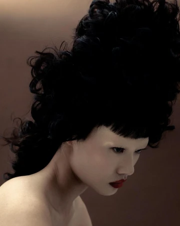 Studio shot of a fashionably made-up pale-skinned woman with dark red lipstick and an elaborate updo, set against a gradient brown background.