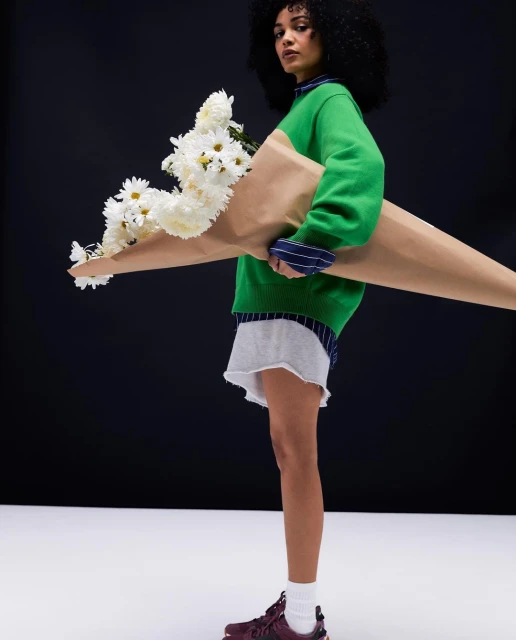 Young adult woman in a green sweater carries a large bouquet of white daisies against a dark background, wearing a gray skirt and maroon sneakers.