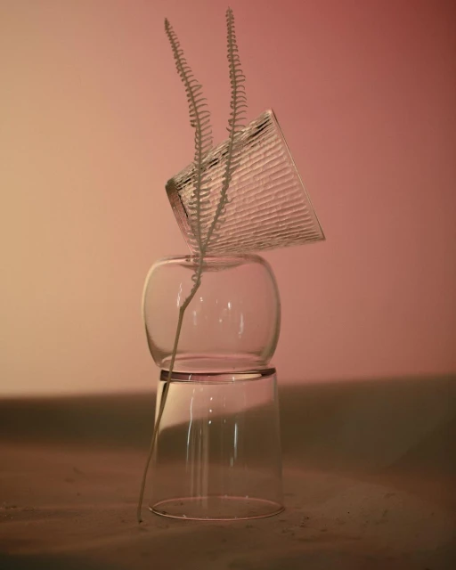 Still life of three clear glass cups stacked vertically with a single sprig of greenery on a gradient background, creating a minimal aesthetic.