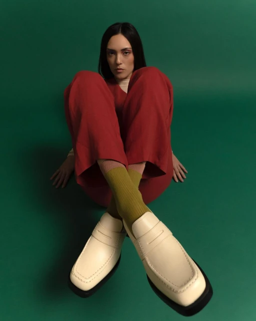 Portrait of a young adult woman sitting indoors, wearing red pants and cream-colored loafers, against a green backdrop.