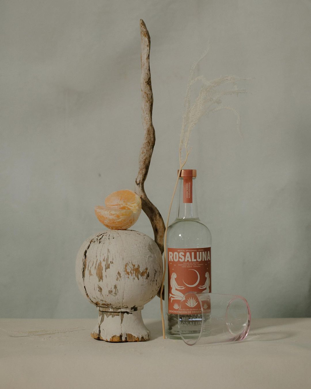 Still life composition featuring a Rosaluna bottle, orange peel, wooden stand, and branch against a muted blue textured background.