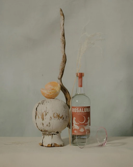 Still life composition featuring a Rosaluna bottle, orange peel, wooden stand, and branch against a muted blue textured background.