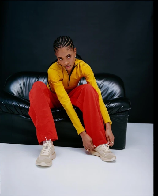 A woman sitting on a black leather couch, tying the laces of her beige sneakers. She's wearing a yellow shirt and red pants.