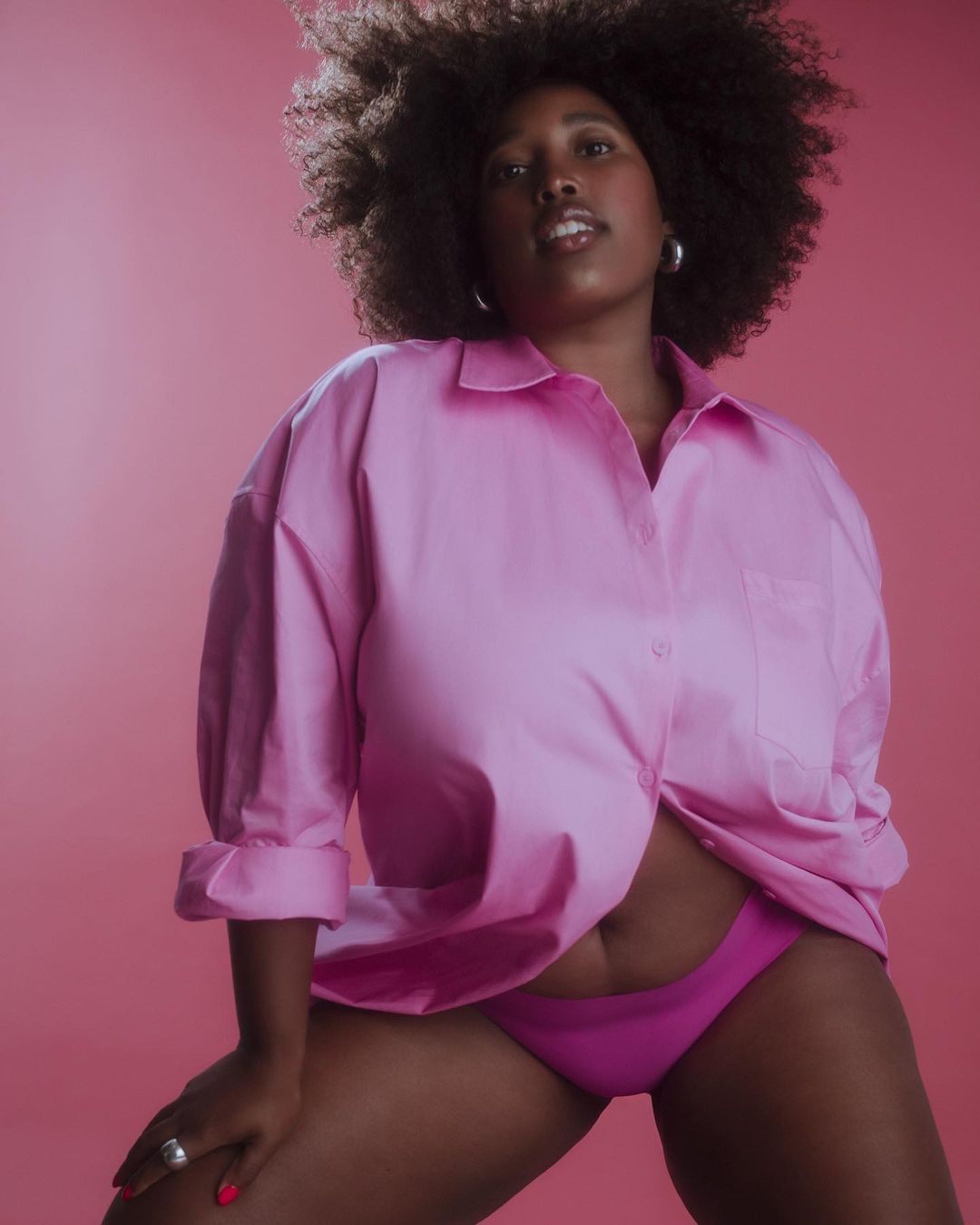 Portrait of an adult African-descent woman with afro hairstyle, wearing a pink unbuttoned shirt and pink underwear against pink background.