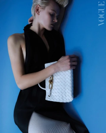 Blonde woman wearing a black halter top holding a white woven leather handbag against a bright blue studio wall.